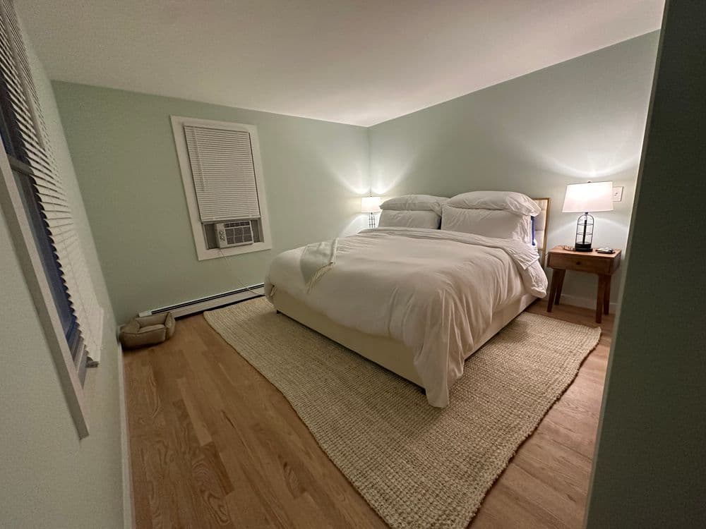 Cozy bedroom with a queen bed, nightstand, and air conditioner on a light green wall.