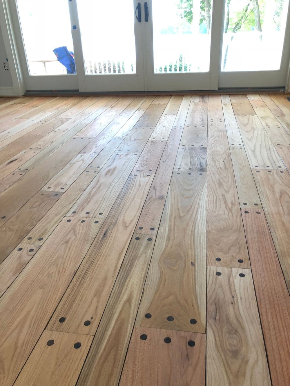 Wood flooring installation with visible nail patterns, natural light from large windows.