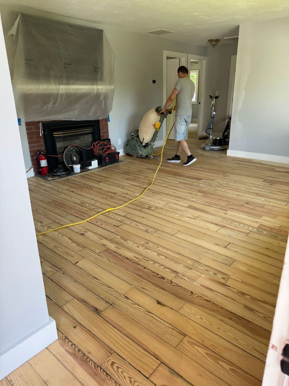 Man refinishing hardwood floors in a bright, renovated living room with tools and equipment.